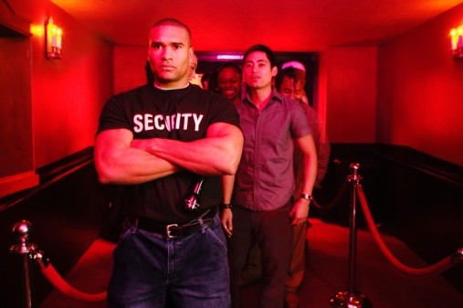 African male bouncer with arms crossed in front of line of people
